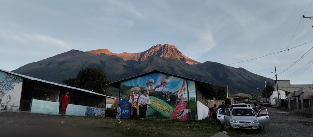 A town street with a mural-covered building beneath a sunlit mountain.
